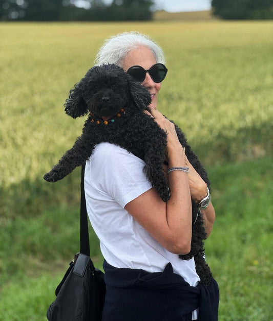 Schwarzer Zwergpudel Rocca auf dem Arm von Adrea, mitten im Park. Beide wirken glücklich und entspannt. Rocca trägt eine Romero Bernsteinkette von ecopetique.